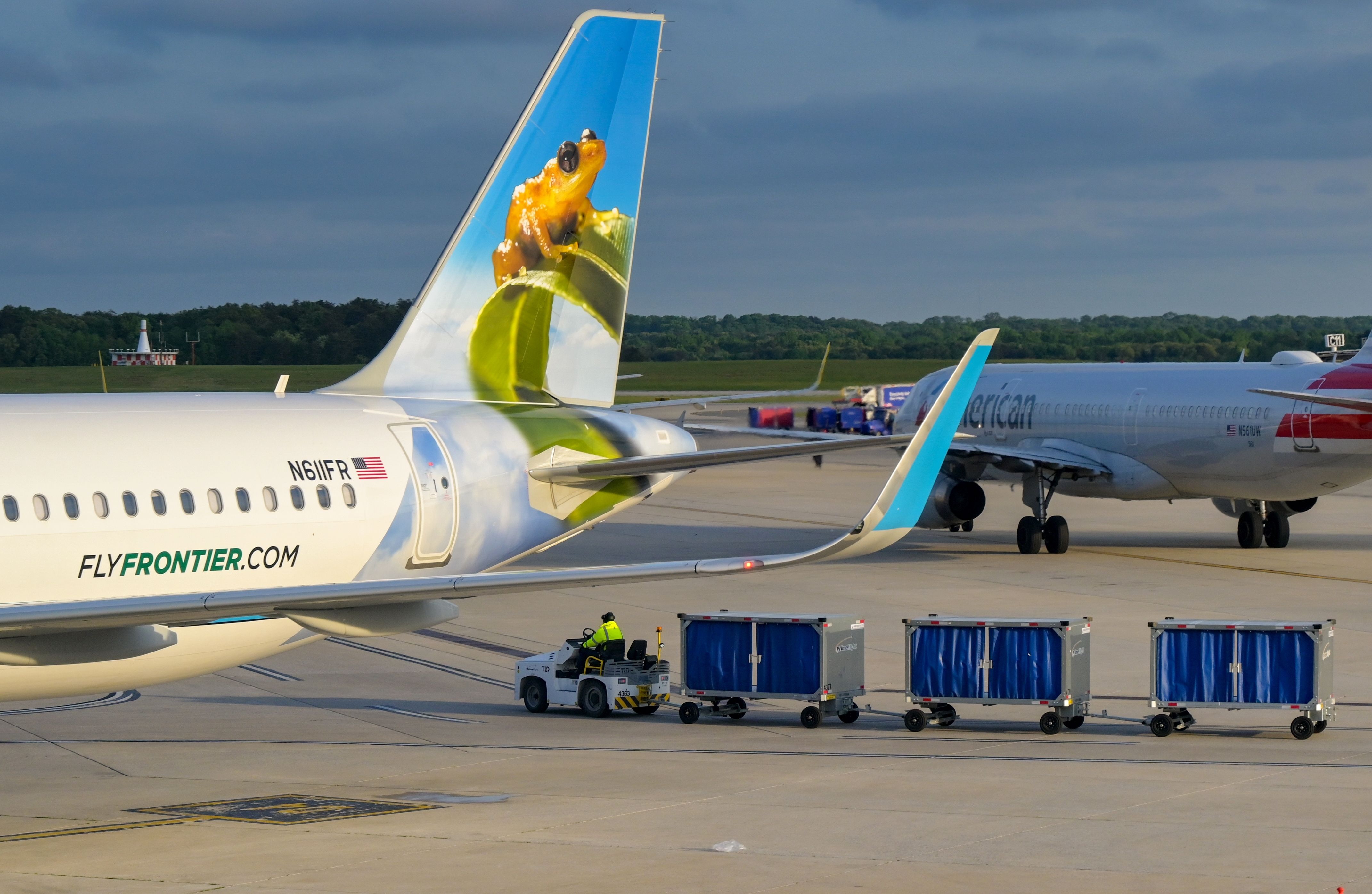Tail fin of Airbus A320 jet operated by Frontier Airlines (registration N611FR) at Baltimore Washington International airport with luggage carts being towed and an American Airlines plane in the background