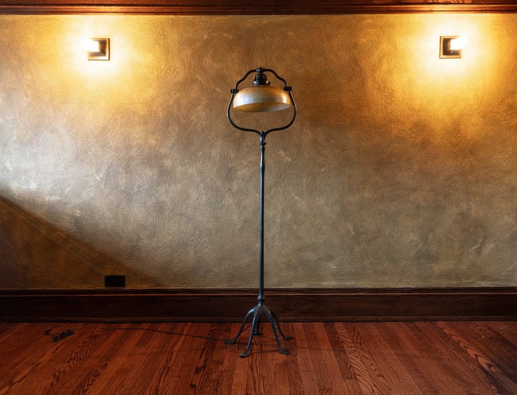 Tall metal floor lamp with sculptural base against textured wall and sconces.