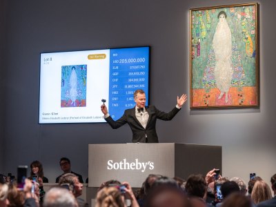 A man in a tuxedo with both arms raised, a painting of a woman hangs behind him with on the screen to its left reads the current bid for the painting in multiple currencies. 