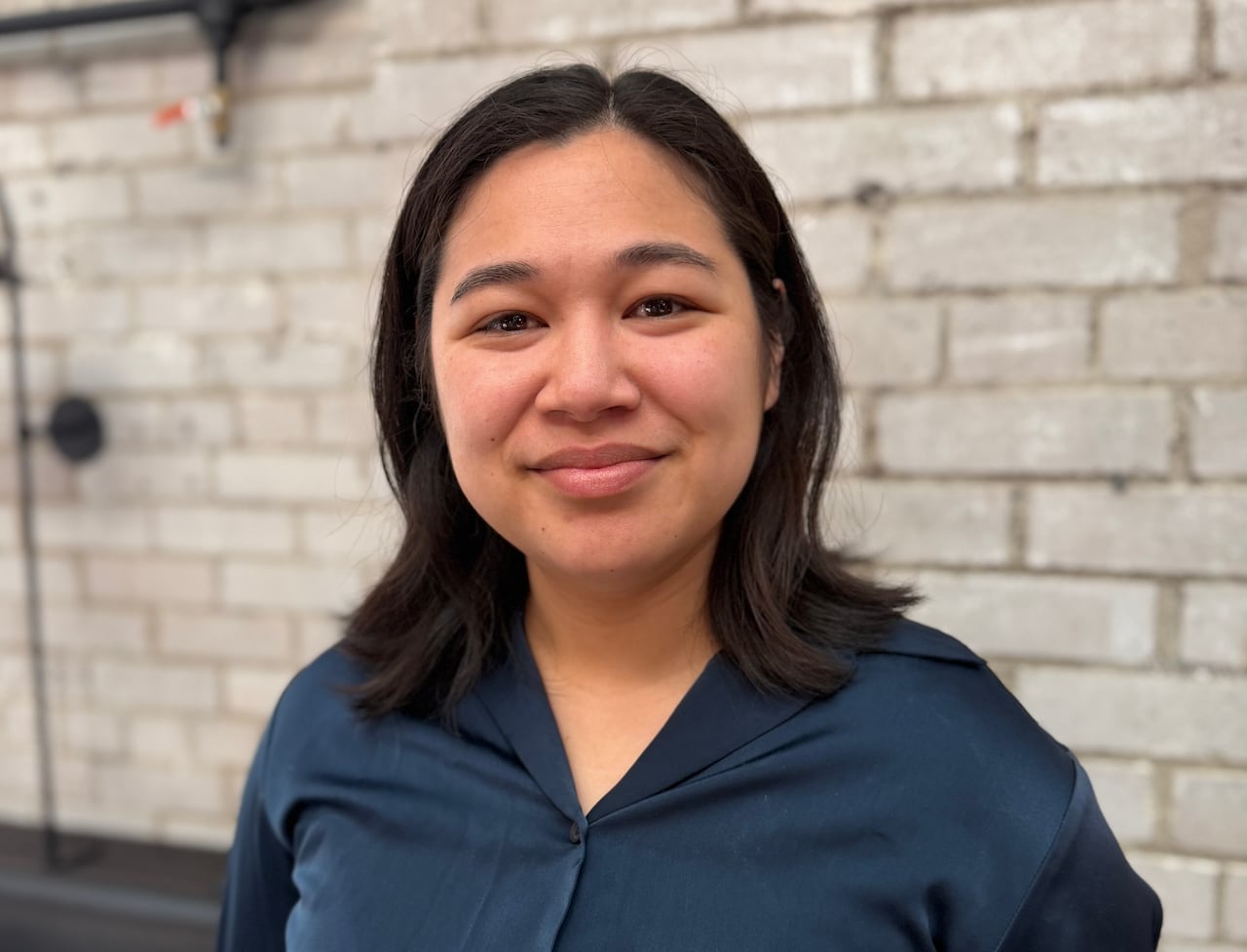 Woman with dark hair wearing blue shirt smiles at camera with a white brick background behind her.