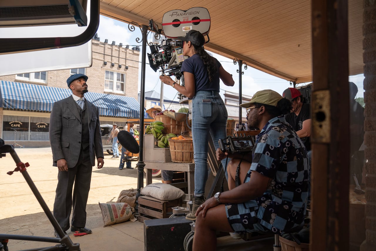 A man wearing a suit and a blue hat looks up at a camera operated by woman wearing a cap with her hair in a long braid. A man in a white hat wearing a blue checked shirt and shorts sits behind her looking at a video monitor.