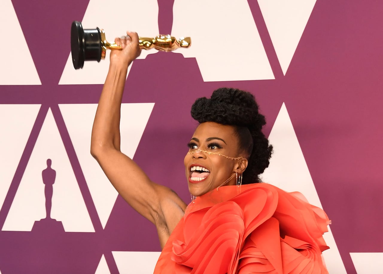Woman wearing red-orange ruffled dress holding Oscar statue in the air. 