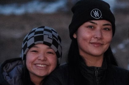 Woman wearing checkered toque smiling and standing next to woman wearing Westworld toque. See part of a camera in her hand.