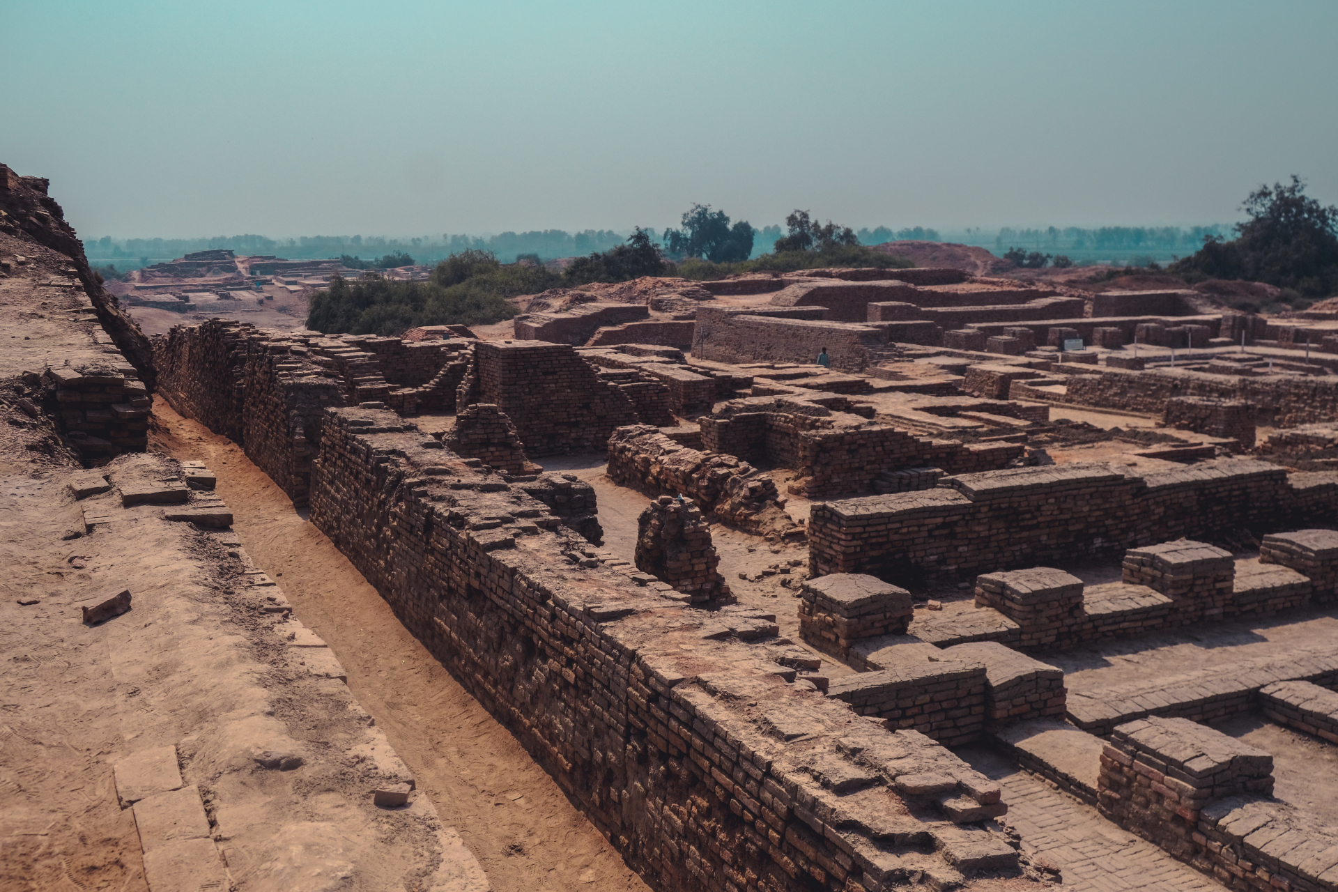 A crumbling city has brown brick walls and dried roads with trees in the background
