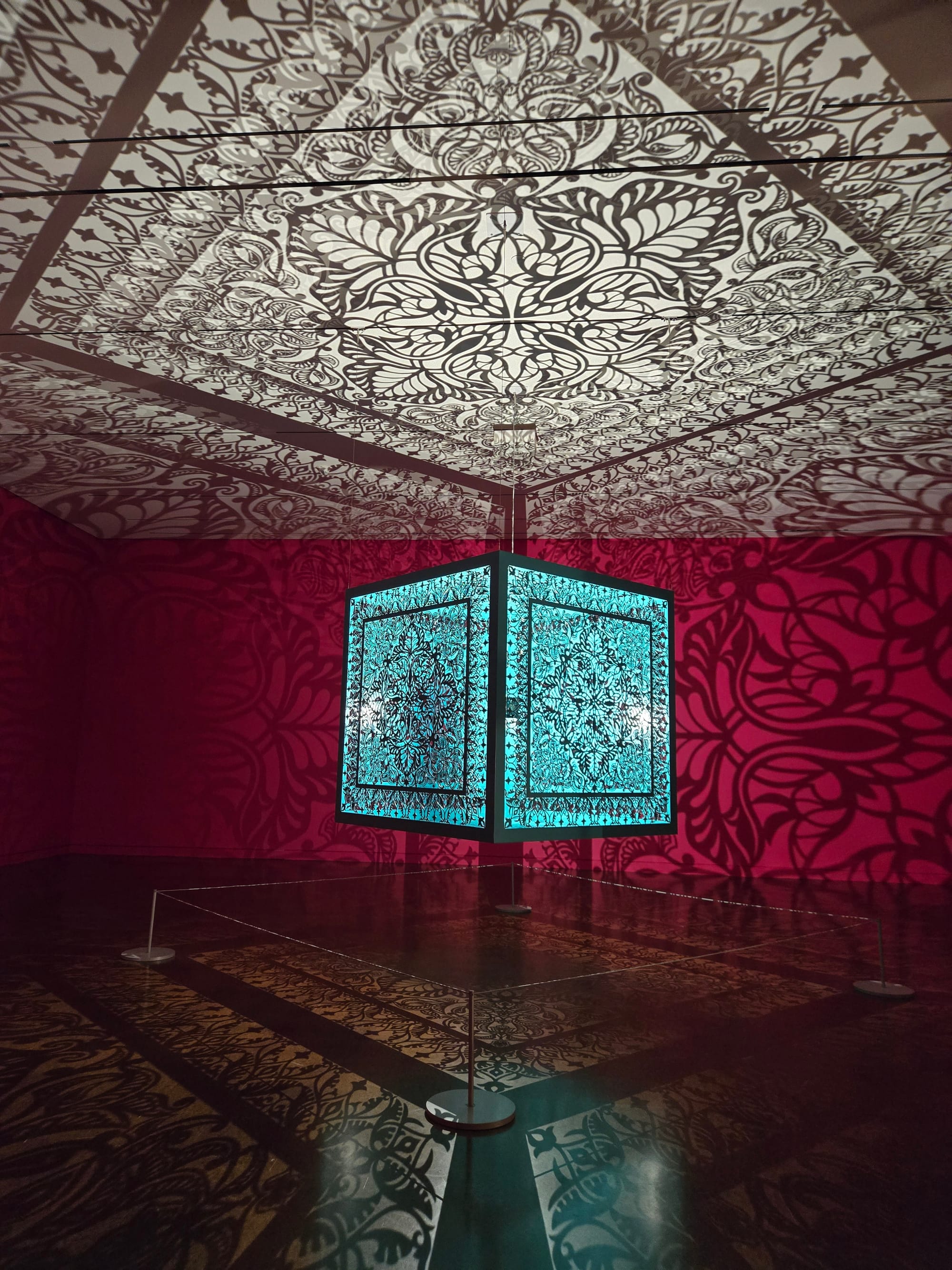 A blue cube hangs in the middle of an empty room with red walls, a black floor and white ceiling. Cutouts in the cube project intricate patterns on all the room's surfaces.