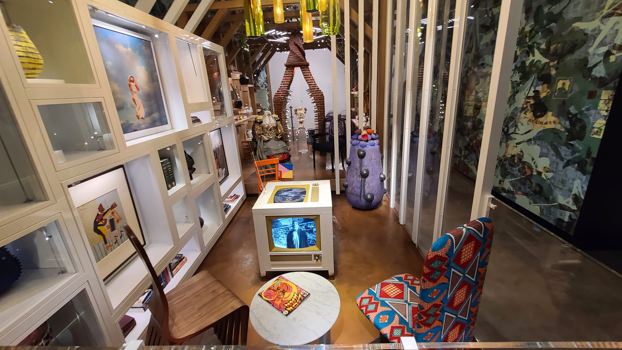 A museum exhibition with white shelving and framed art on the left side, two chairs on the floor, an old-fashioned television in the middle of the room. A purple sculpture in the back underneath a chandelier made of green and yellow glass, and a brick open sculptural chimney in the background.