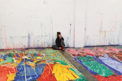 An artist seated on the floor of her studio, leaning against a wall, with large-scale designs on the floor in front of her.