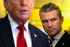 A man with slicked back hair stands behind another older man as he speaks.