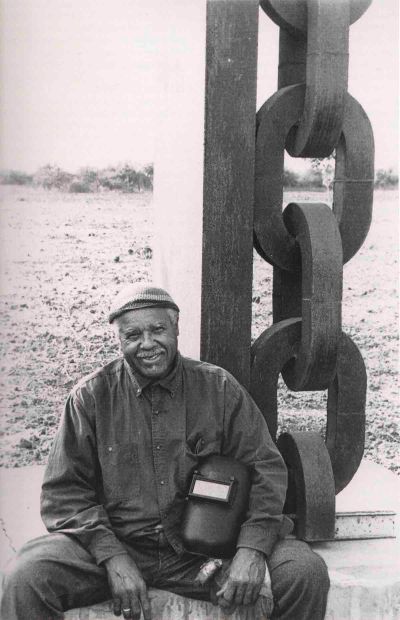 A man with a welder's helmet seated near a large sculpture of a chain.