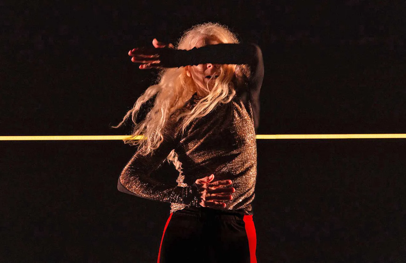 A woman with long blonde hair and a sparkly gold shirt shielding her face with her arm, standing against a dark background with a yellow horizontal light bar.