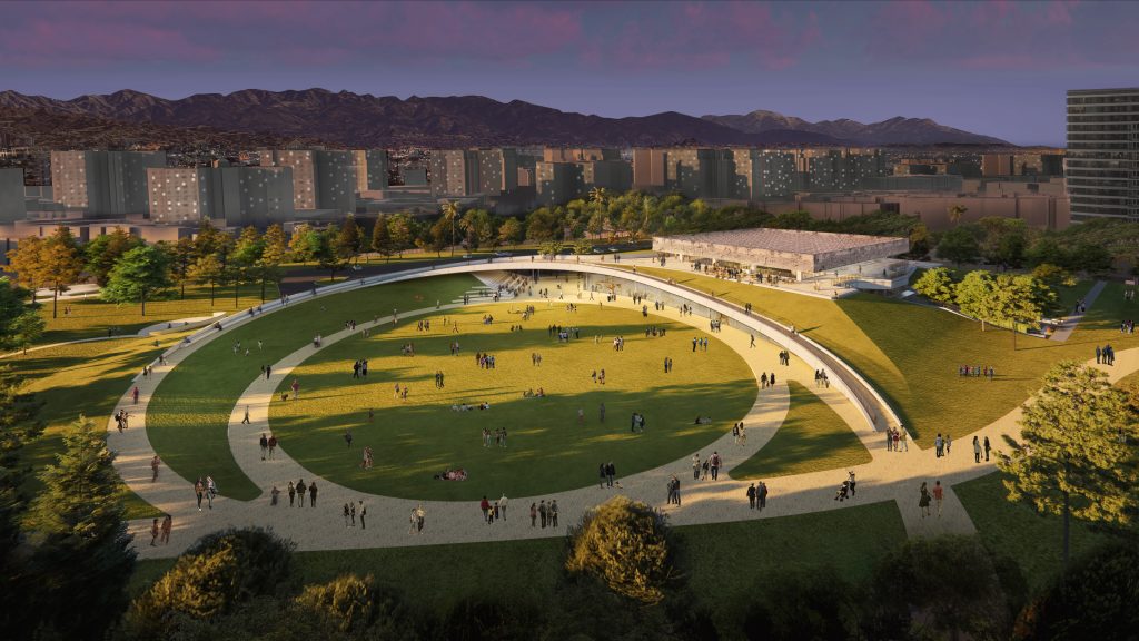 An architectural rendering of a white, single story museum built into a hill overlooking a green field surounded by circular walking paths, all situated beneath a sunset sky with the mountains of L.A. looming over off in the distance. The campus will open before the Olympic games