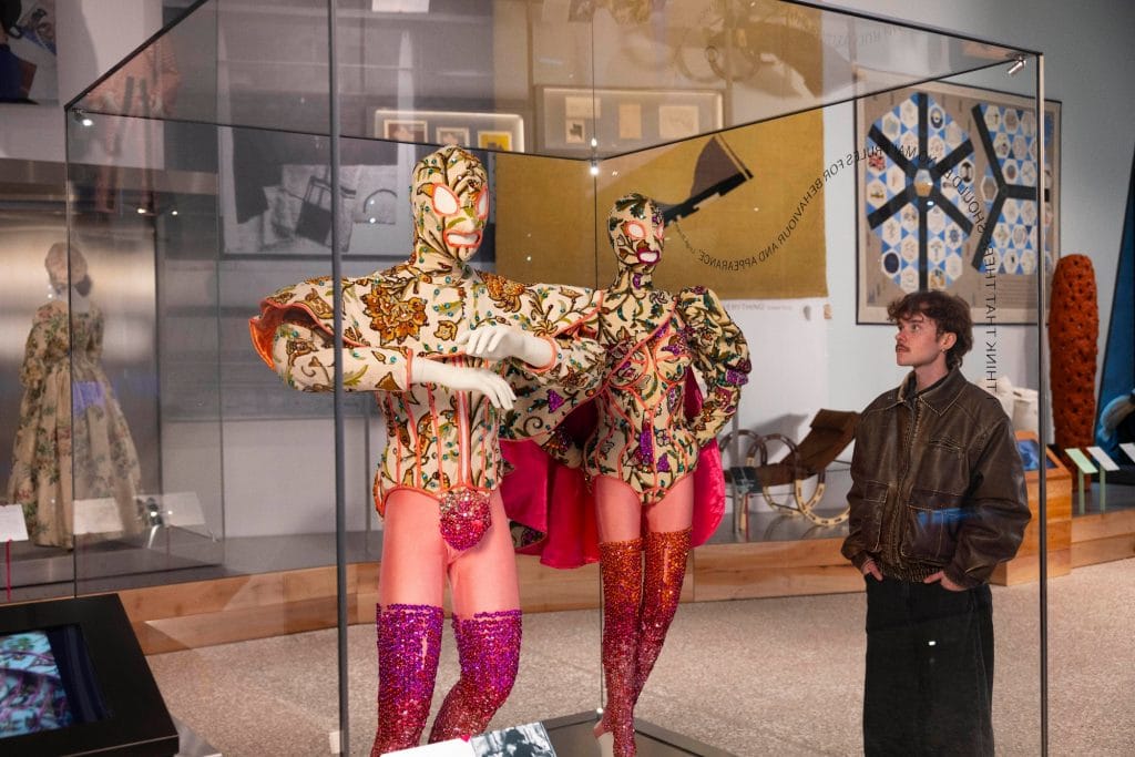 two elaborate pieces of pink-hued fancy dress are on mannequins in a glass museum cabinet