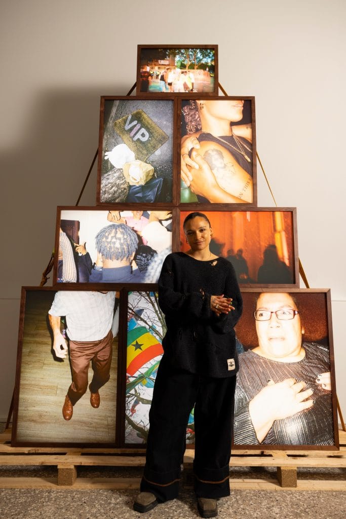 a person dressed in black stands in front of a sculptura piece composed of photographs piled on top of each other in a triangle formation