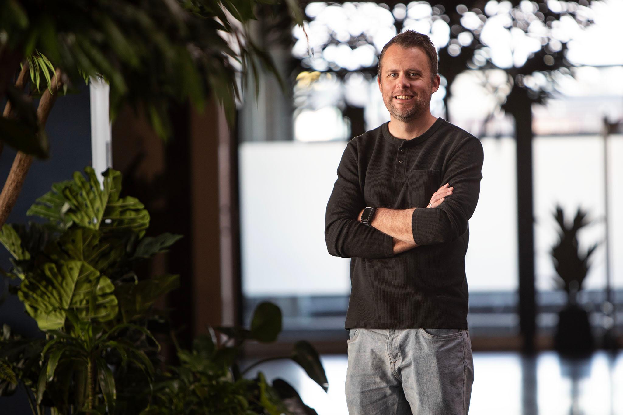 A man in a black shirt crosses his arms and looks into the lens, in a room of large, bright windows and potted plants.
