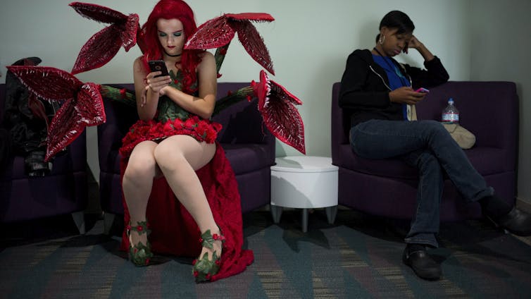 A person in a red and green costume with plant-like tentacles, wearing high heels and lipstick, sits in a chair looking at a phone.