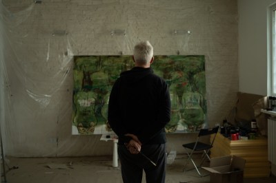 A man, with his back to the camera, looks at a painting in-progress in his studio. He holds a paintbrush. 