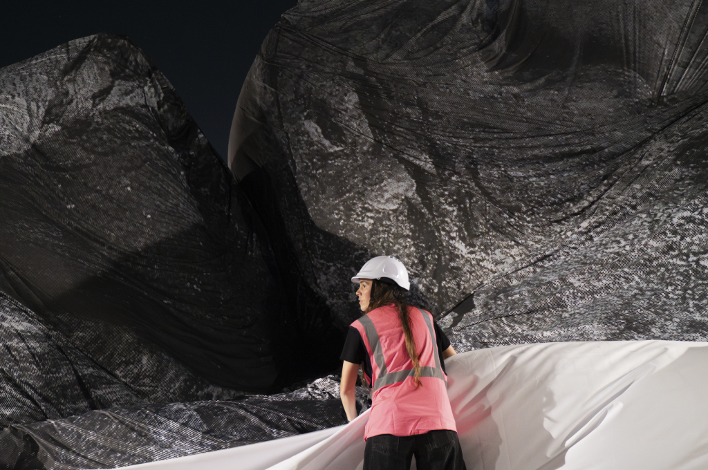 A photograph of a girl in high visibility vest and white hard hat helpingn to hold up a large piece of fabric amidst an inflatable tunnel