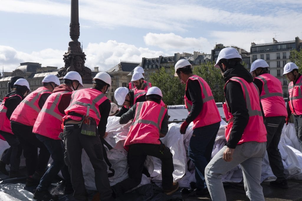 A crowd of twelve people in pink high visibility vests and white hard hats heaves fabric on the Pont Neuf bridge in Paris on a partly cloudy afternoon