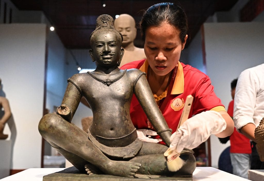 Museum conservator carefully cleans seated bronze Khmer statue from Cambodia while examining intricate surface details