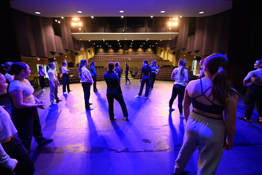 A large group of Saint Anselm College dancers  stand onstage in a proscenium theater.