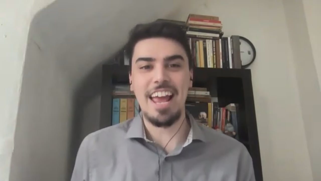 A man in a grey shirt sits in front of a bookshelf.