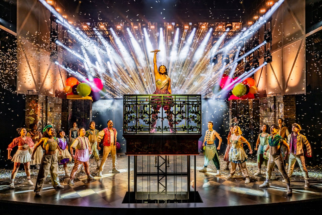 A woman stands on a small balcony with her fist in the air and fireworks behind her, while a group of brightly-dressed people stand on the ground looking up at her.
