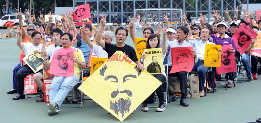 A photograph of a crowd of seated people bearing photos and protest posters mostly featuring the face of Ai Weiwei