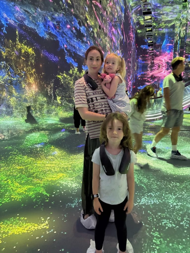 A woman holds a young child while taking a mirror selfie inside a vivid immersive art installation with colorful projected biological imagery on walls and floor