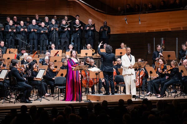 An orchestra performs onstage in a concert hall, in front of a chorus. Two singers stand in front of the orchestra.