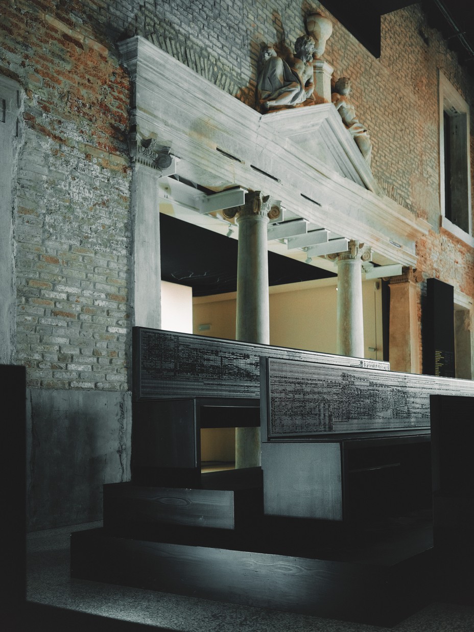 photo of installation with dark stair-stepped wooden benches in large brick hall with marble pediment and sculptures over wide doorway with marble columns