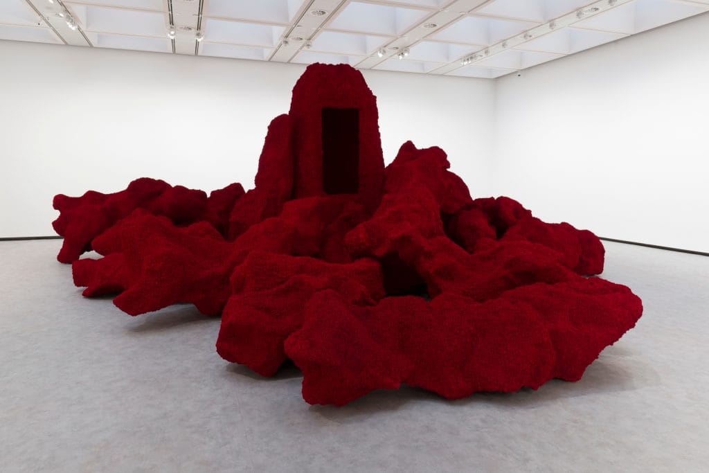 a very large red artwork in a white walled room, it looks almost like a rocky terrain with a small hut emerging out of it 
