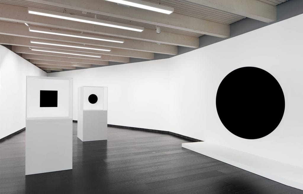 small black artworks are mounted on white podiums in a white walled space, on the right a black circle hovers on the wall