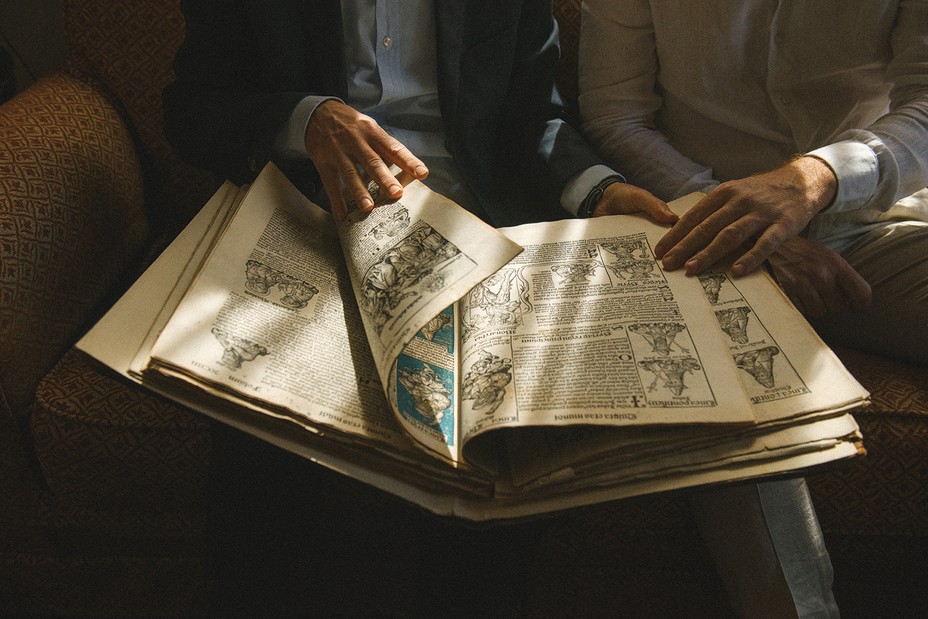 photo of very large illustrated manuscript as two men's hands page through it