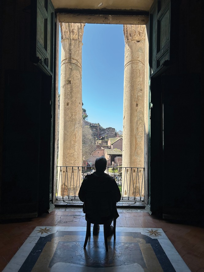 photo from behind of man sitting on chair facing extremely tall doorway open onto columned portico with railing and view of bright blue sky and Roman ruins