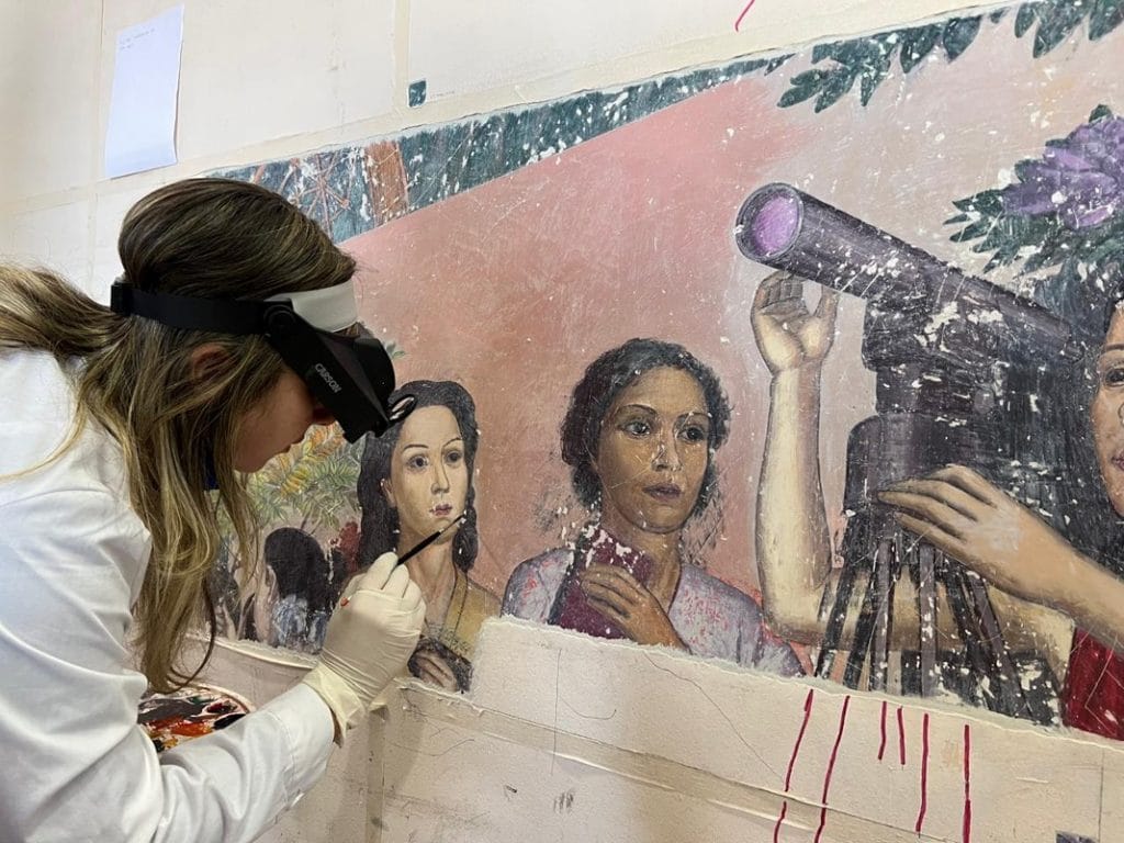 Conservator carefully retouches uncovered Julio Escámez mural while wearing magnifying visor and protective gloves during restoration.