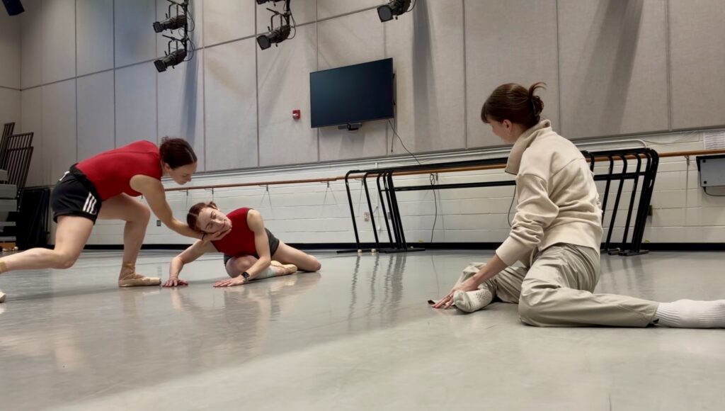 In a studio, a choreographer sits on the floor while watching two dancers rehearse. One lunges forward as the other pushes up on her arms, her head tilted and her legs behind her.