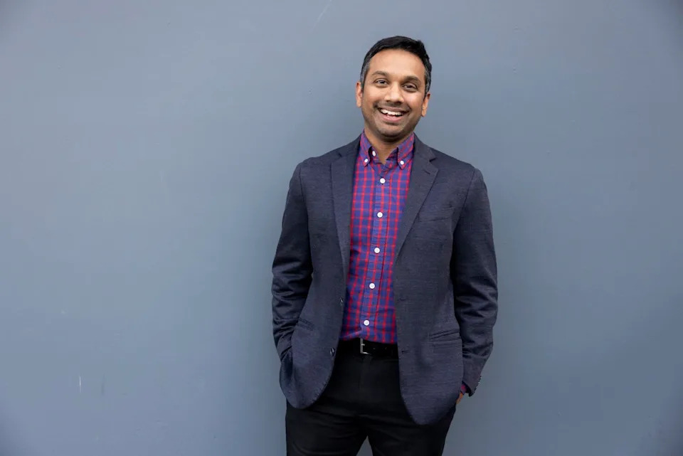 A man smiles in front of a large gray wall.