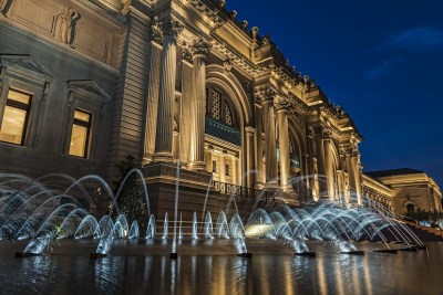 A lit-up building at twilight with a fountain in front of it.