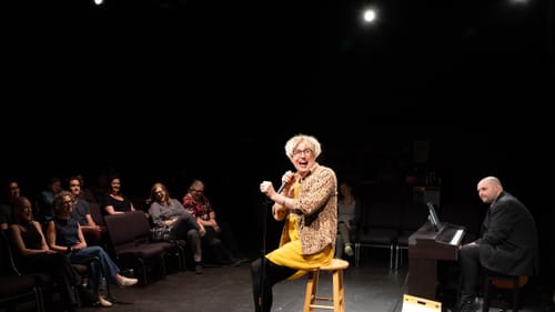Kitrosser, in modest yellow drag with a leopard-print shirt, smiles joyfully at the mic in front of a small theater audience.