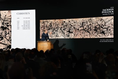 An auctioneer at Christie's stands at the podium, fist raised, selling Jackson Pollock's "Number 7A, 1948," an oil and enamel on canvas drip painting displayed on a large screen behind him. A currency board to the left shows a hammer price of $157 million in multiple currencies. The silhouettes of a packed salesroom audience fill the foreground.