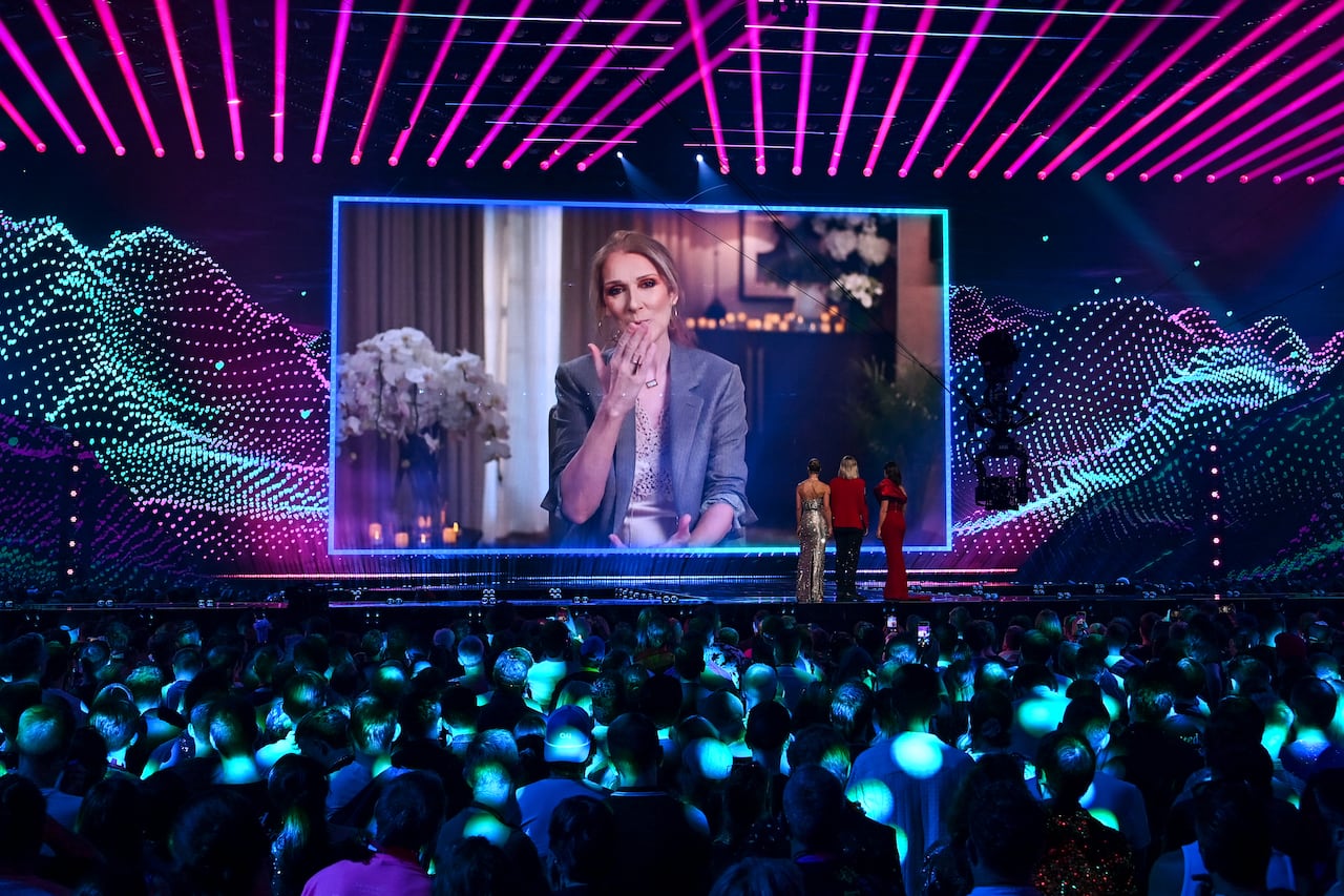 A large crowd of people look at a stage, where three women in evening wear stare a screen. On the screen, a woman is blowing a kiss to the crowd.