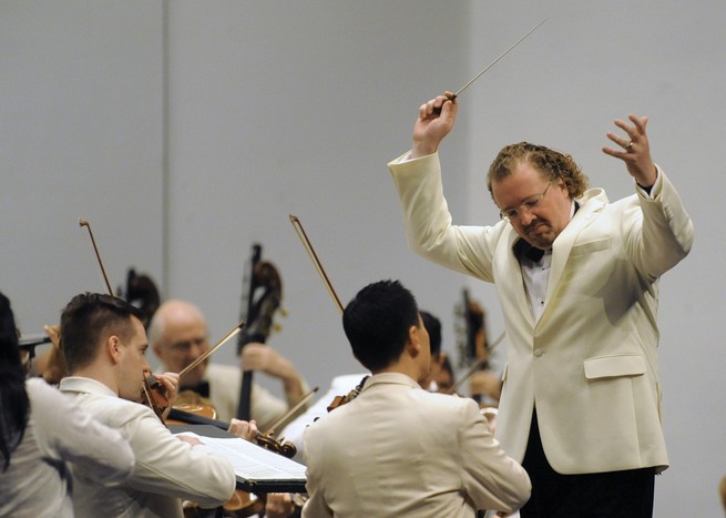 color photo of man in white jacket and black tie and pants with arms raised and eyes closed standing before string section