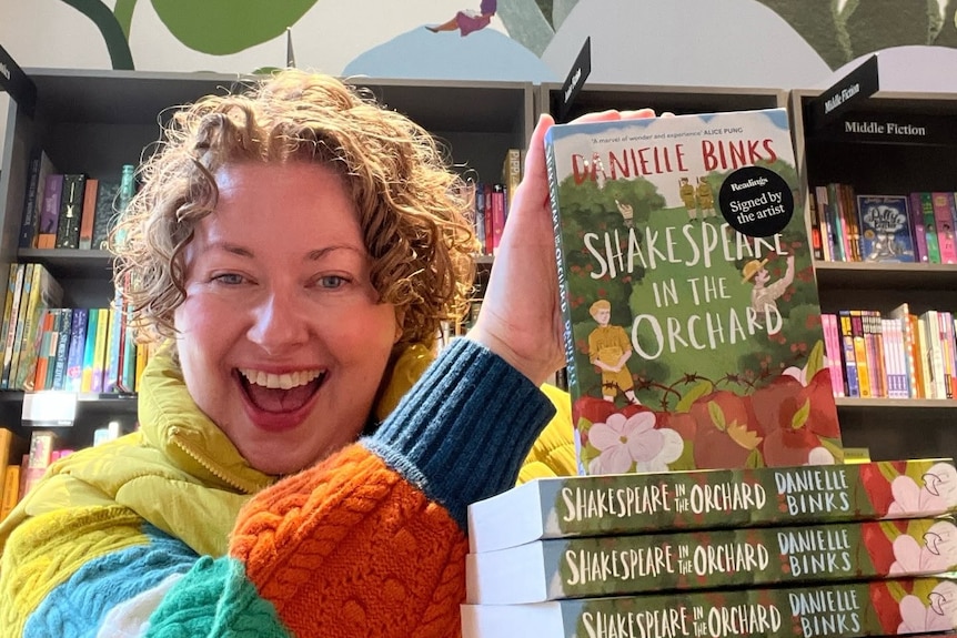 A woman, Danielle Binks, smiles at the camera holding a pile of books recently released