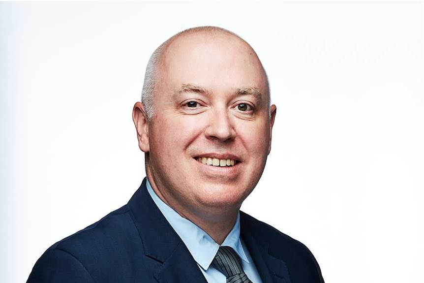 Matthew Warren, a middle aged man in a suit, smiles at the camera in front of a white background