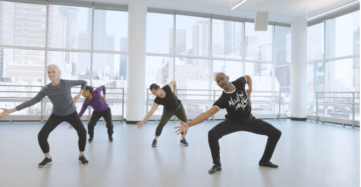 A diverse group of dancers expressing movement in a bright studio with large windows overlooking a city skyline.