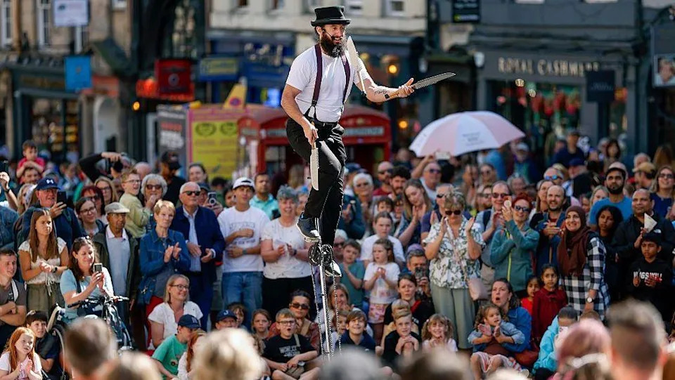 Man on a high unicycle with long knives in his hands, about to thrown them in the air. There is a large crowd watching him on Edinburgh's Royal Mile. He has a beard and is wearing a white T-shirt, black trousers, braces and a black hat.