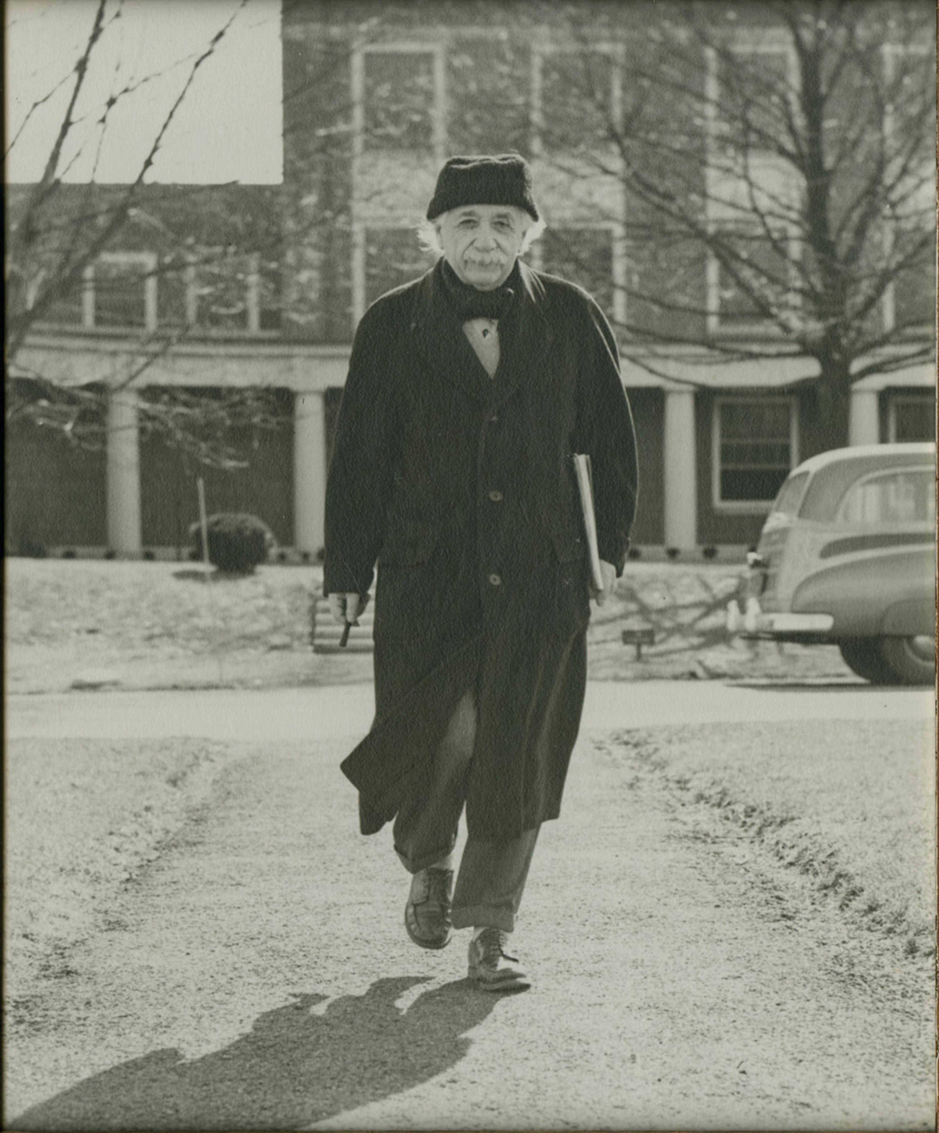 Black-and-white photo of an elderly man in a coat and hat walking down a path with a building and car in the background.