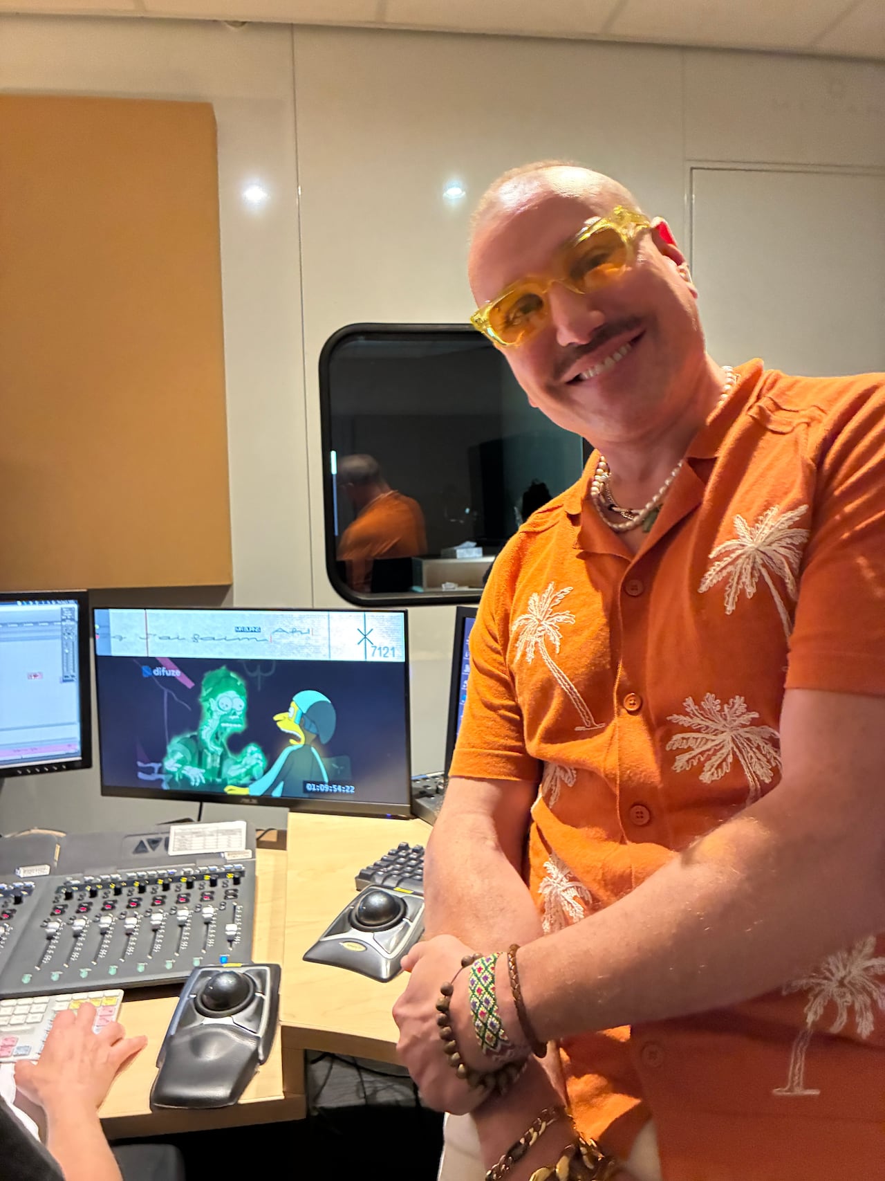 A man wearing an orange button up t-shirt and orange sunglasses smiles at the camera.