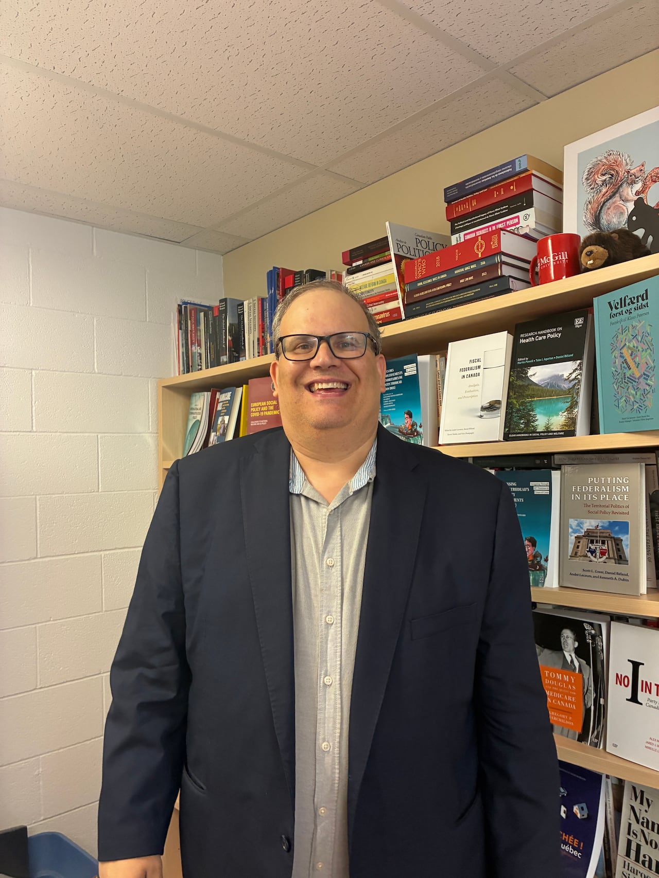 A man wearing a blue suit smiles at the camera with a bookcase behind him.