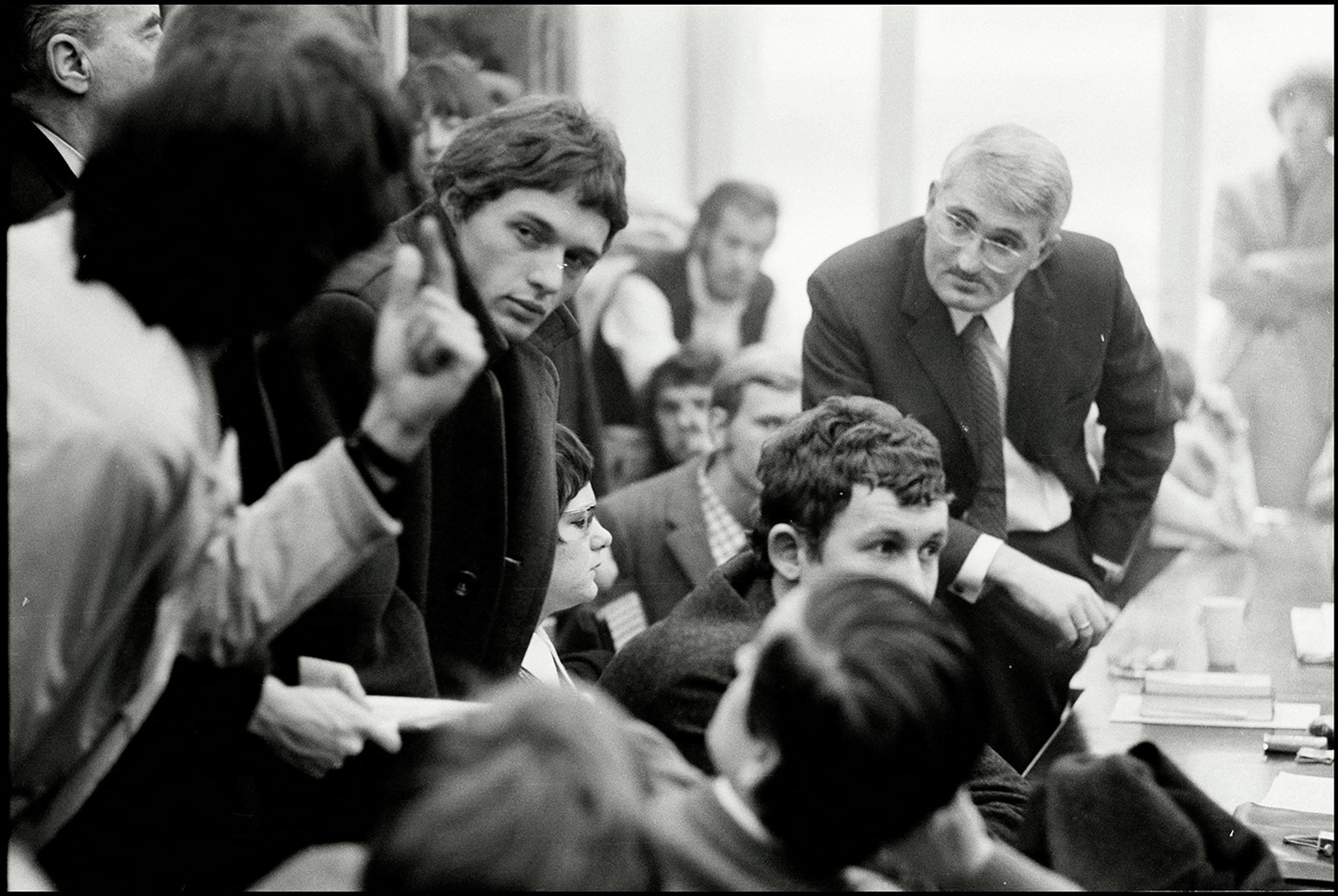 Black-and-white photo of a heated discussion in a crowded room, with a man gesturing while others listen attentively.